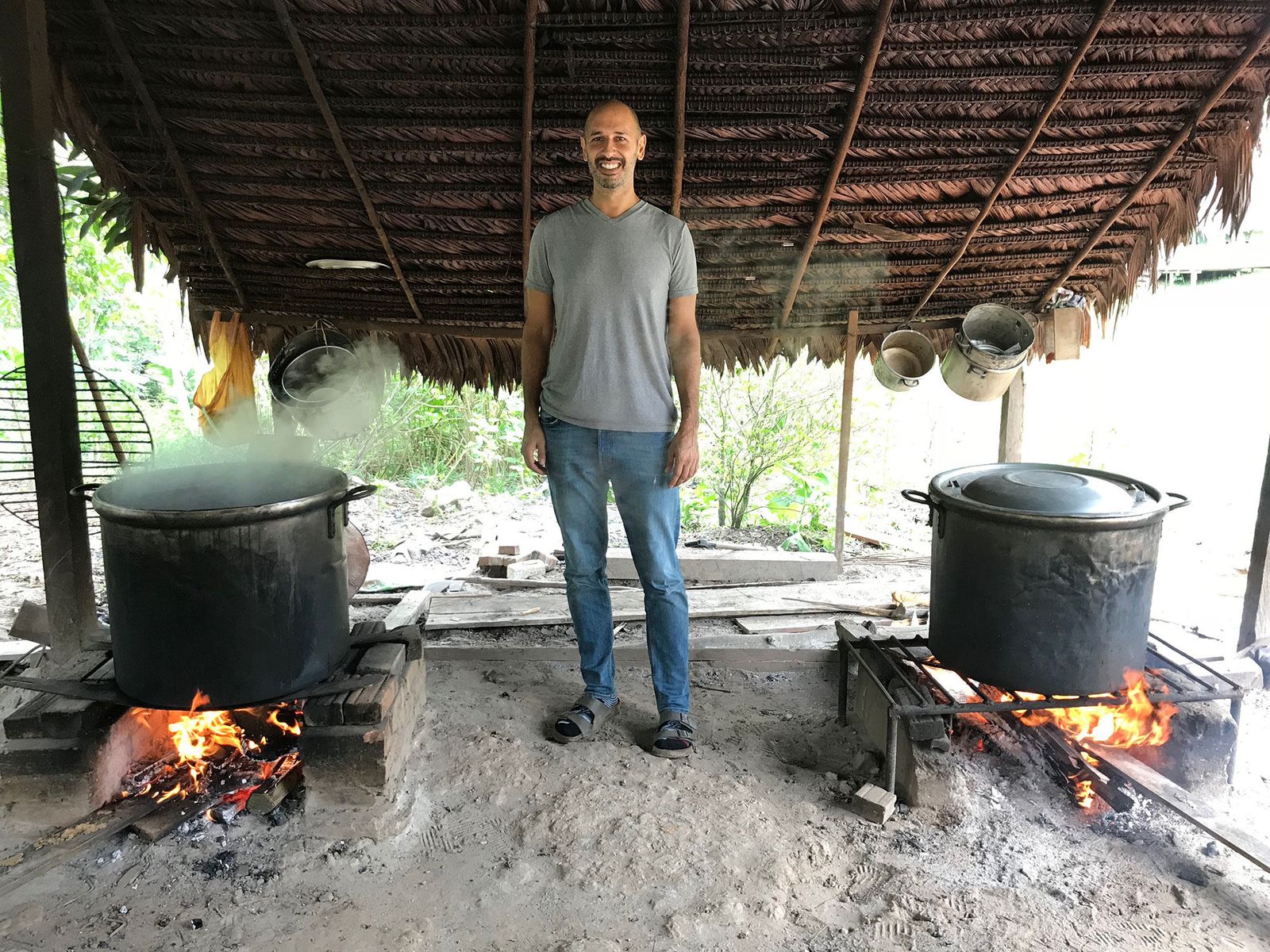 Hamid standing between two large pots of ayahuasca cooking over open fires under a thatched roof, steam rising from one of them