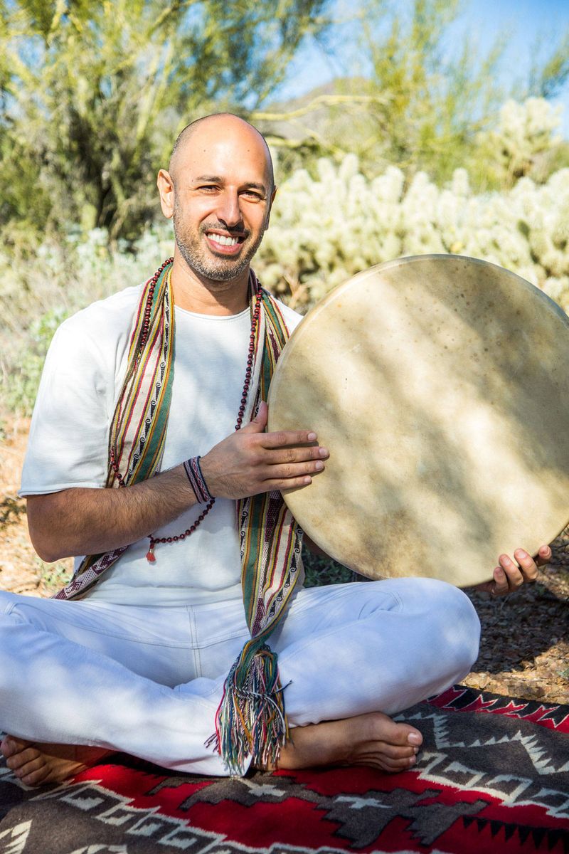 Hamid sitting cross legged outdoors in the desert, smiling, holding a frame drum upright with one hand resting on its skin
