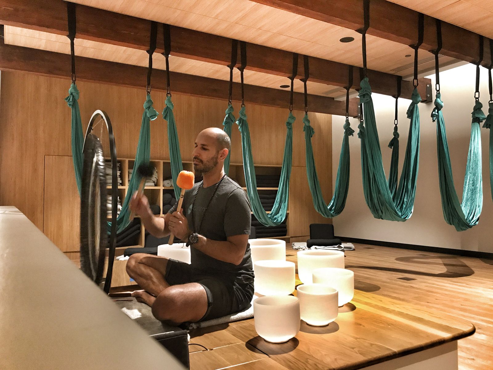 Hamid sitting cross legged on a wooden floor playing a gong with a soft mallet, crystal singing bowls in front of him and teal aerial yoga hammocks hanging from the beams behind