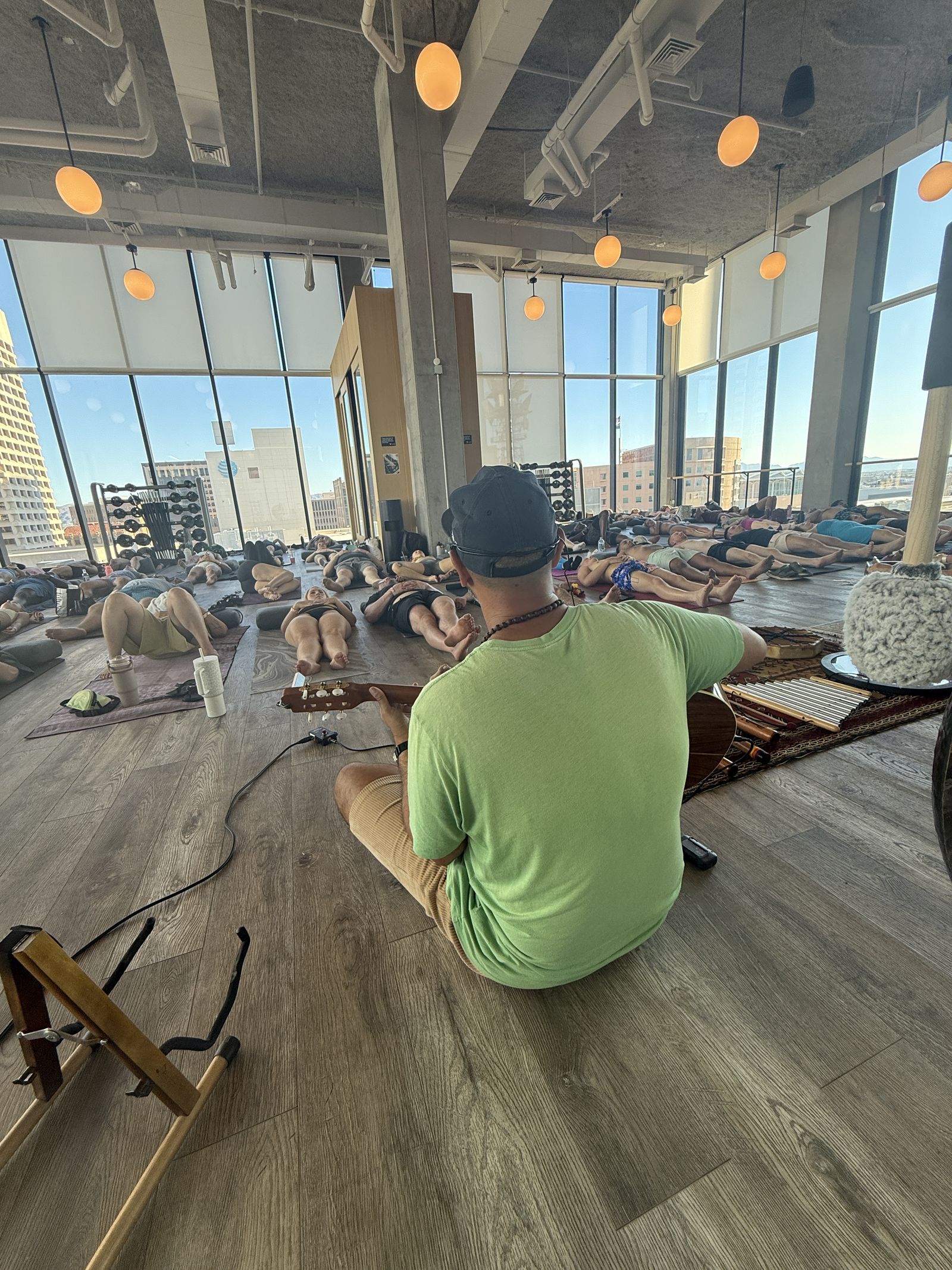 Hamid seen from behind in a green shirt, seated on the floor with his guitar, facing a bright room of people lying on mats before tall windows