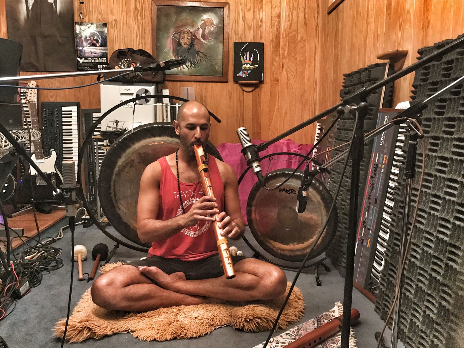 Hamid sitting cross legged on a sheepskin in a recording studio, playing a wooden flute into a microphone, two gongs, keyboards and guitars around him