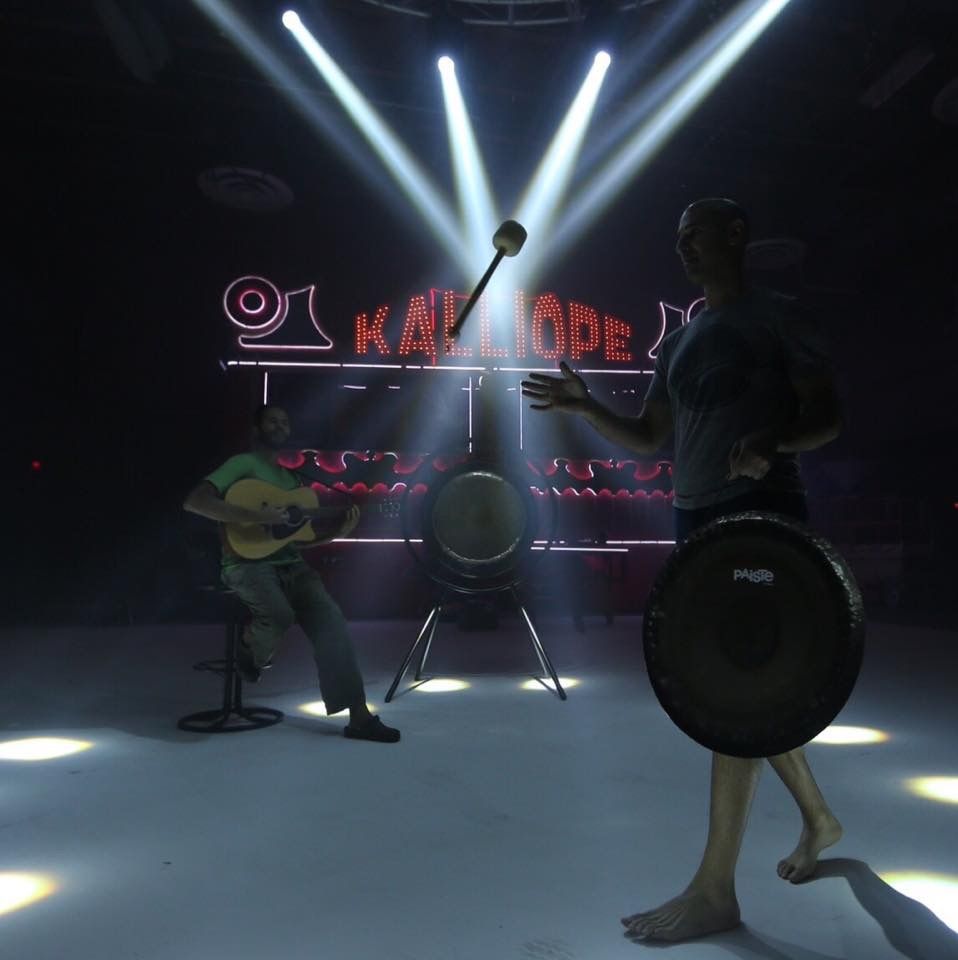 Hamid holding a gong in a dark room under stage lights, a guitarist seated behind him in front of a red neon Kalliope sign