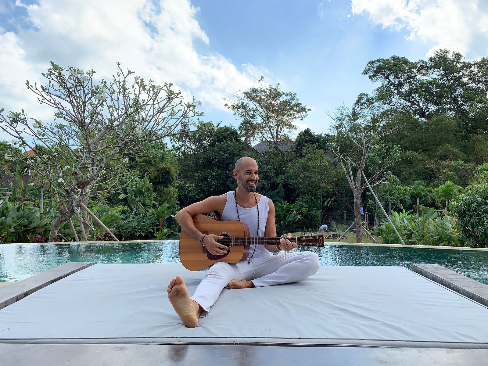 Hamid sitting cross legged beside a pool, playing an acoustic guitar, with dense green trees behind him