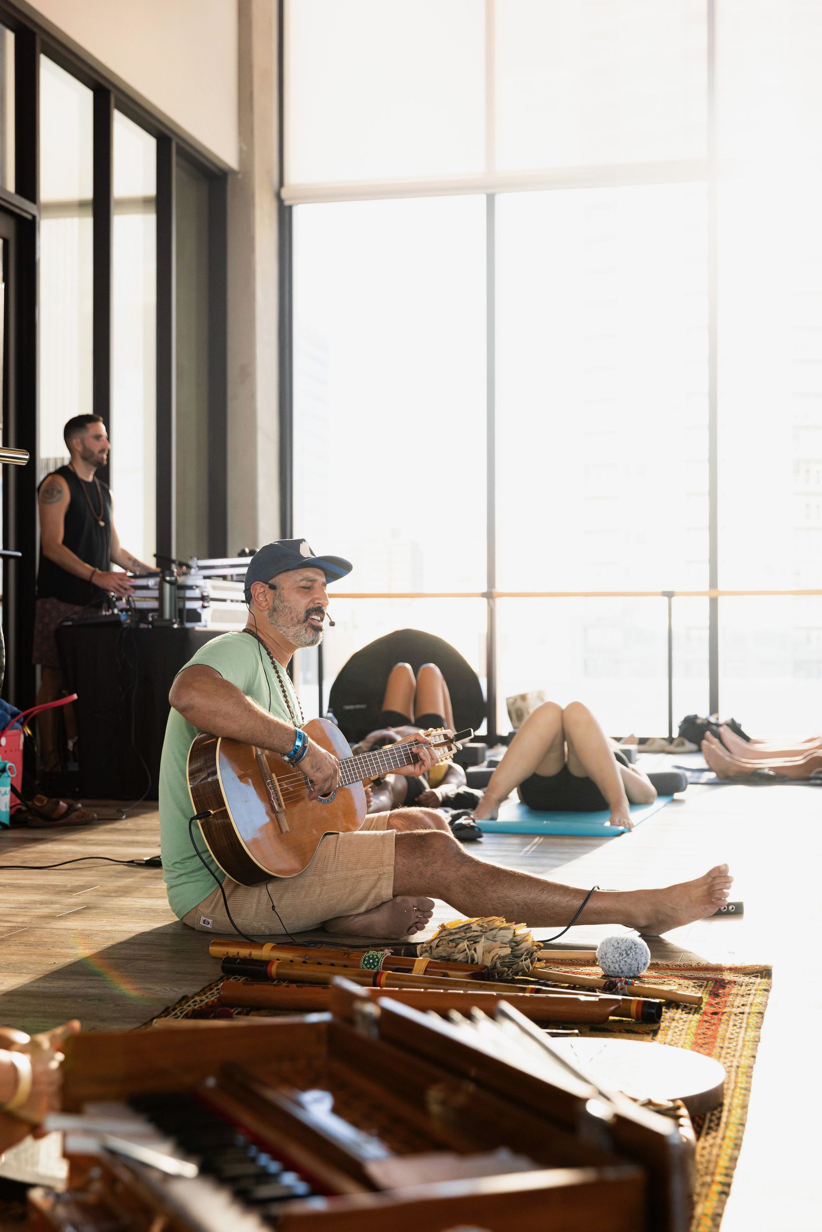 Hamid in a green shirt and cap, seated on the floor playing guitar in window light, a harmonium in front of him and people resting on mats nearby