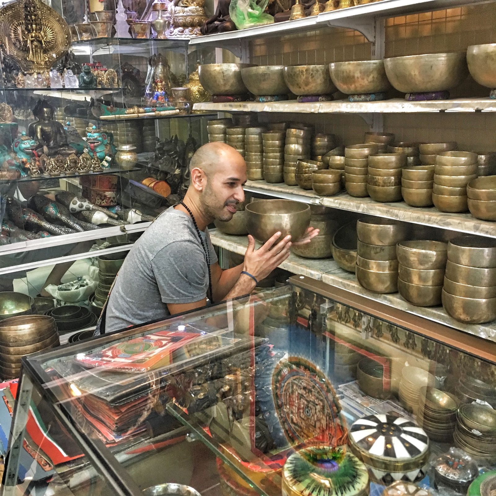 Hamid leaning on a glass counter in a shop lined with shelves of hammered metal singing bowls, holding one of them up in his hands