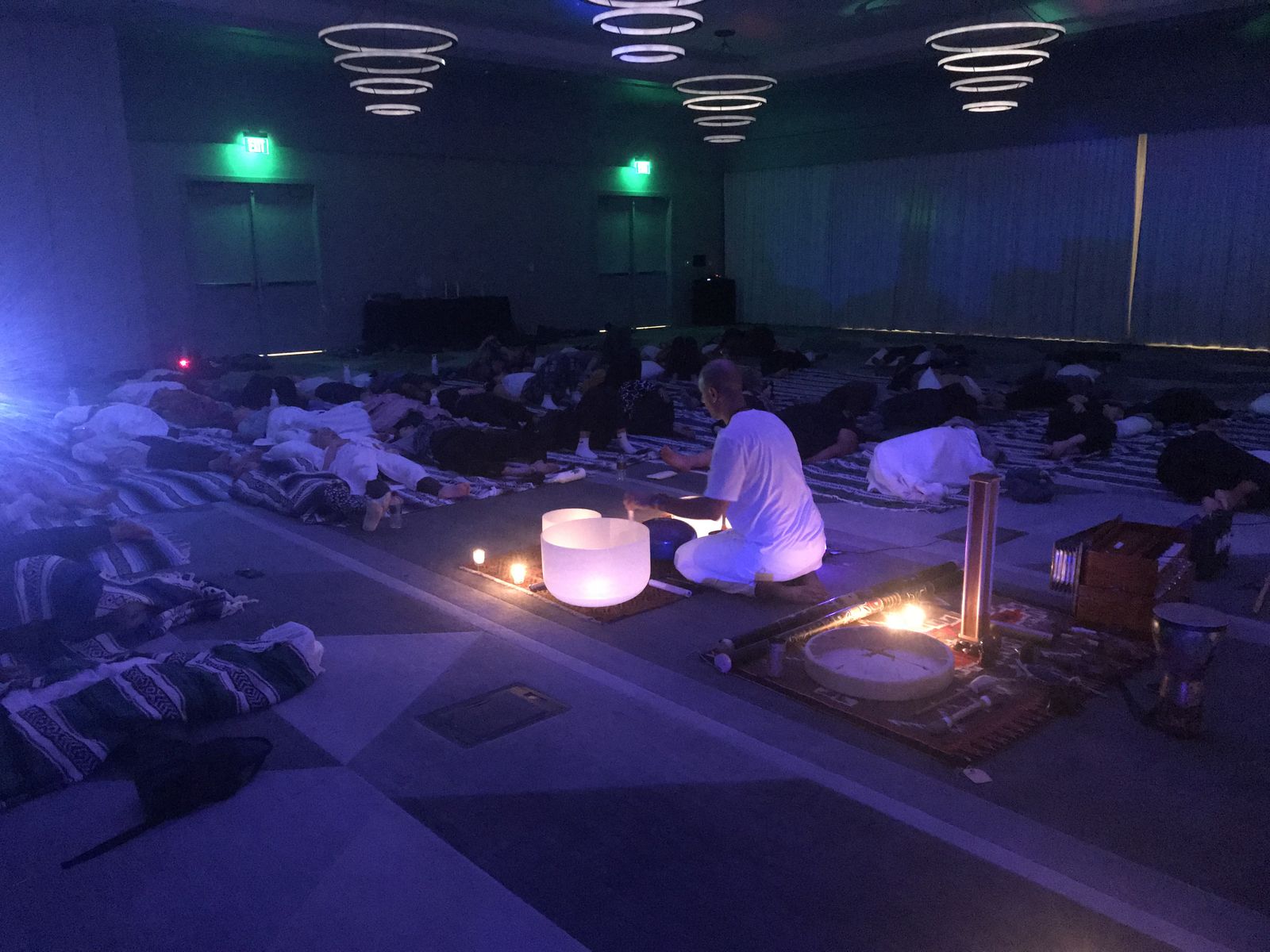 A darkened hotel ballroom under ring chandeliers, rows of people lying on mats, Hamid in white seated among candles beside a large bowl