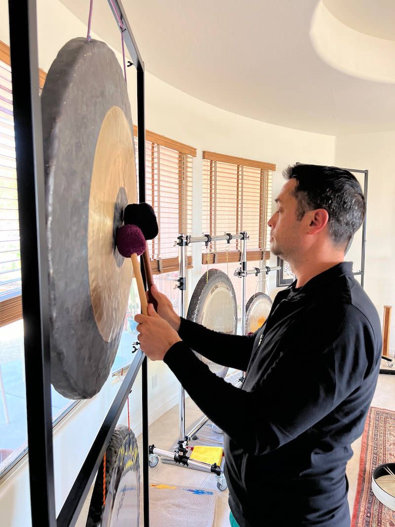 A student at the training playing a large hanging gong with a soft mallet, with three more gongs on stands behind