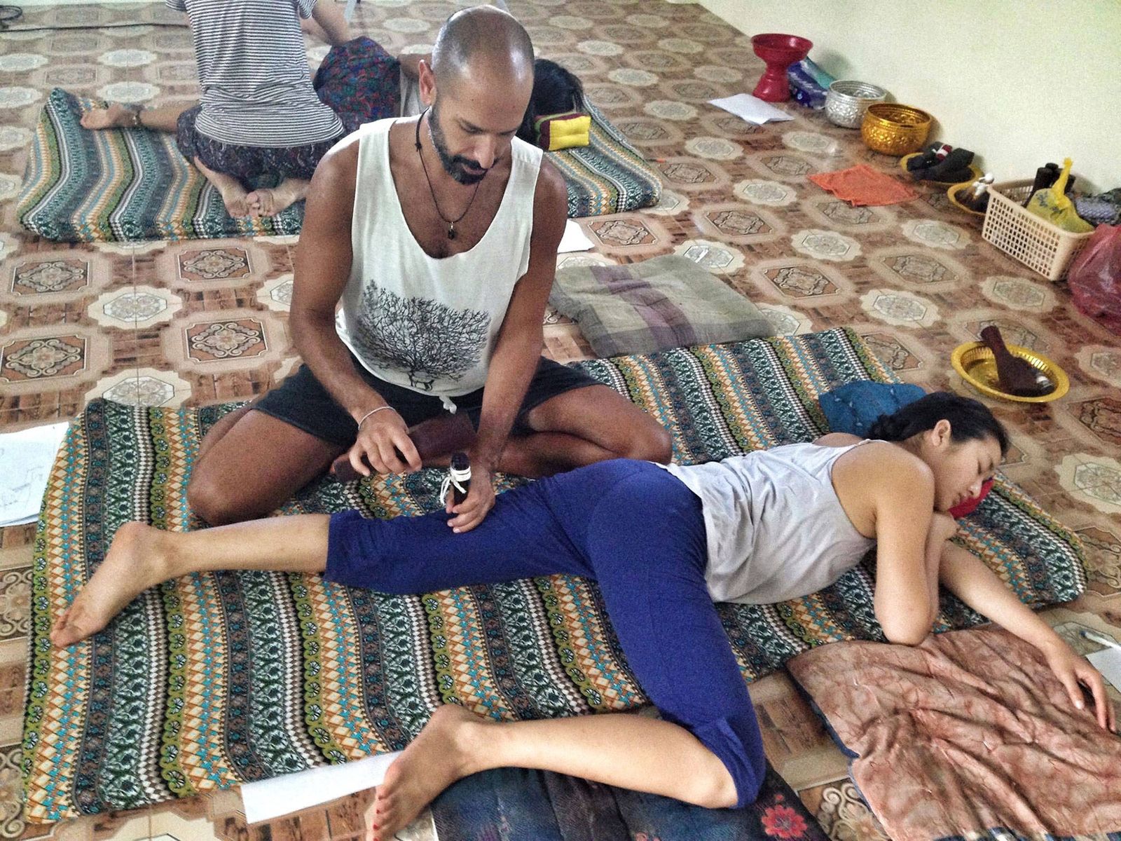 Hamid seated on a patterned mat, working the leg of a woman lying on her side with a wooden peg and mallet, other students on mats behind them