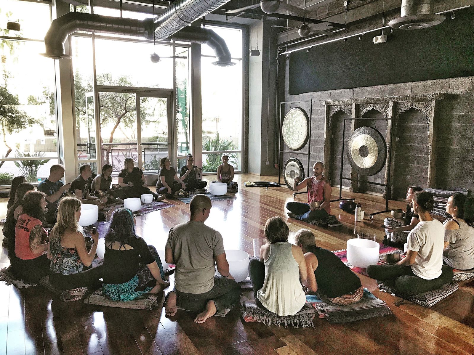 A training circle in a studio, people sitting on mats around crystal bowls, gongs on stands along a brick wall and daylight through tall windows