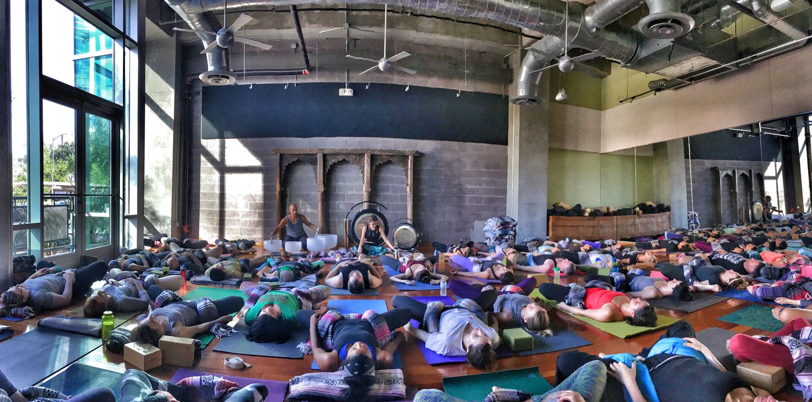 A wide studio room with carved wooden doors and gongs along the back wall, the floor full of people lying on mats