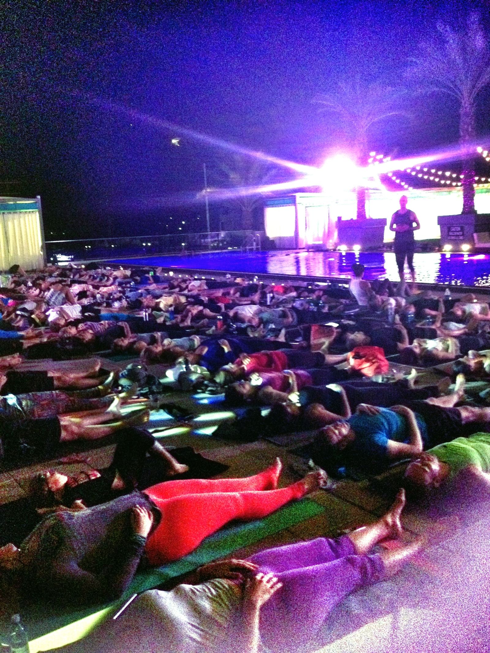 Night on a resort pool deck, a large group lying on mats beside the lit pool, palm trees and a small stage glowing purple behind them
