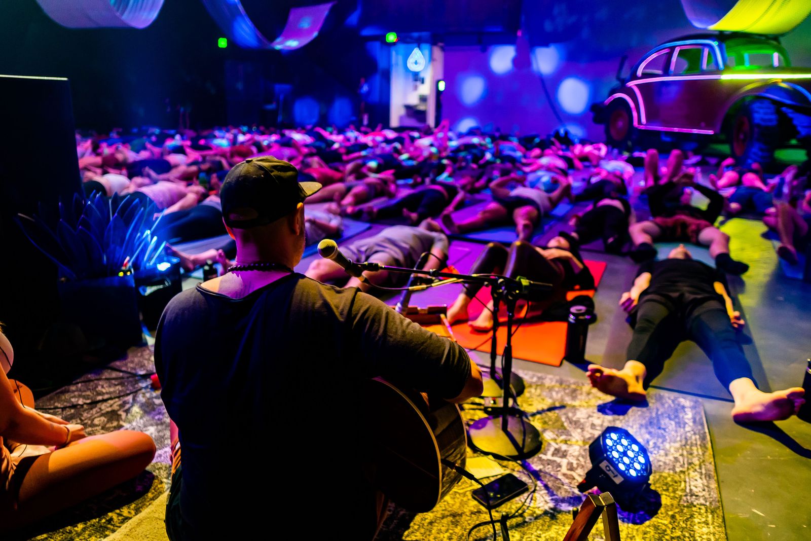 Hamid seen from behind, playing guitar on a rug, facing a room of people lying on mats in blue and pink light