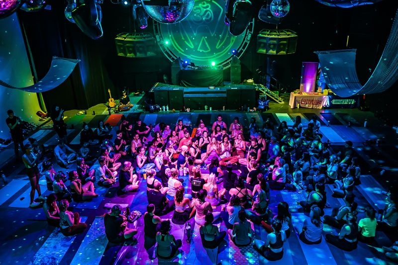 A dark concert hall full of people seated on mats in pink and blue light, mirror balls overhead and a round lit stage behind them, at Walter Studios