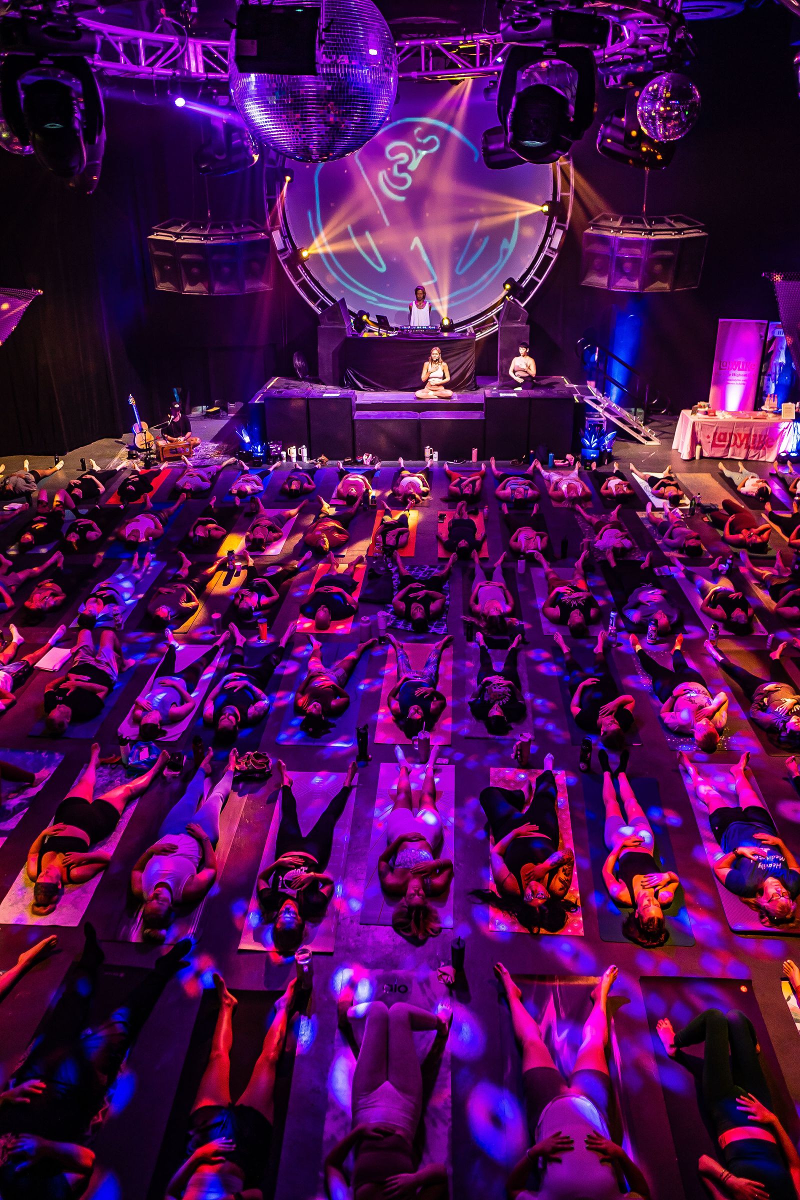 Seen from above, a dark hall filled with rows of people lying on mats under purple stage lights, a large lit mandala over the stage
