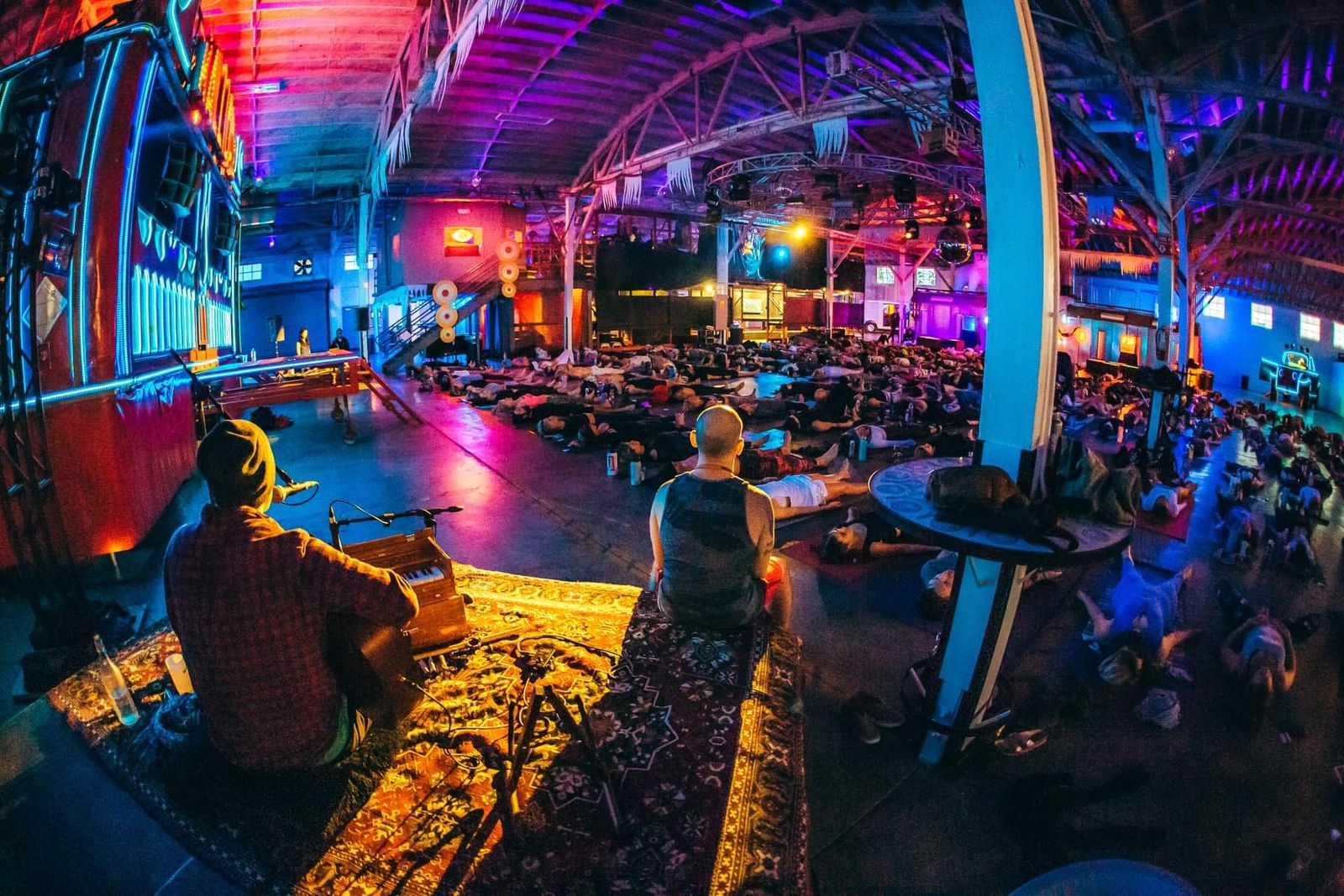 Hamid at a harmonium on a rug beside the stage, facing a warehouse floor of people lying down in colored light