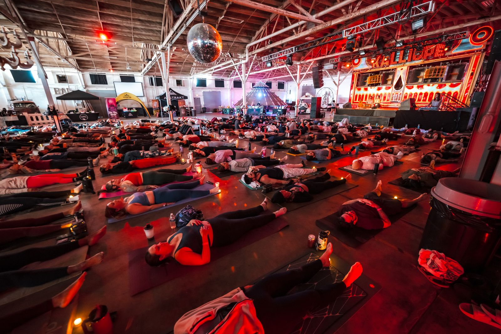A long warehouse floor under a steel truss roof, filled with people lying on mats in red light, candles along the rows, the Kalliope stage behind