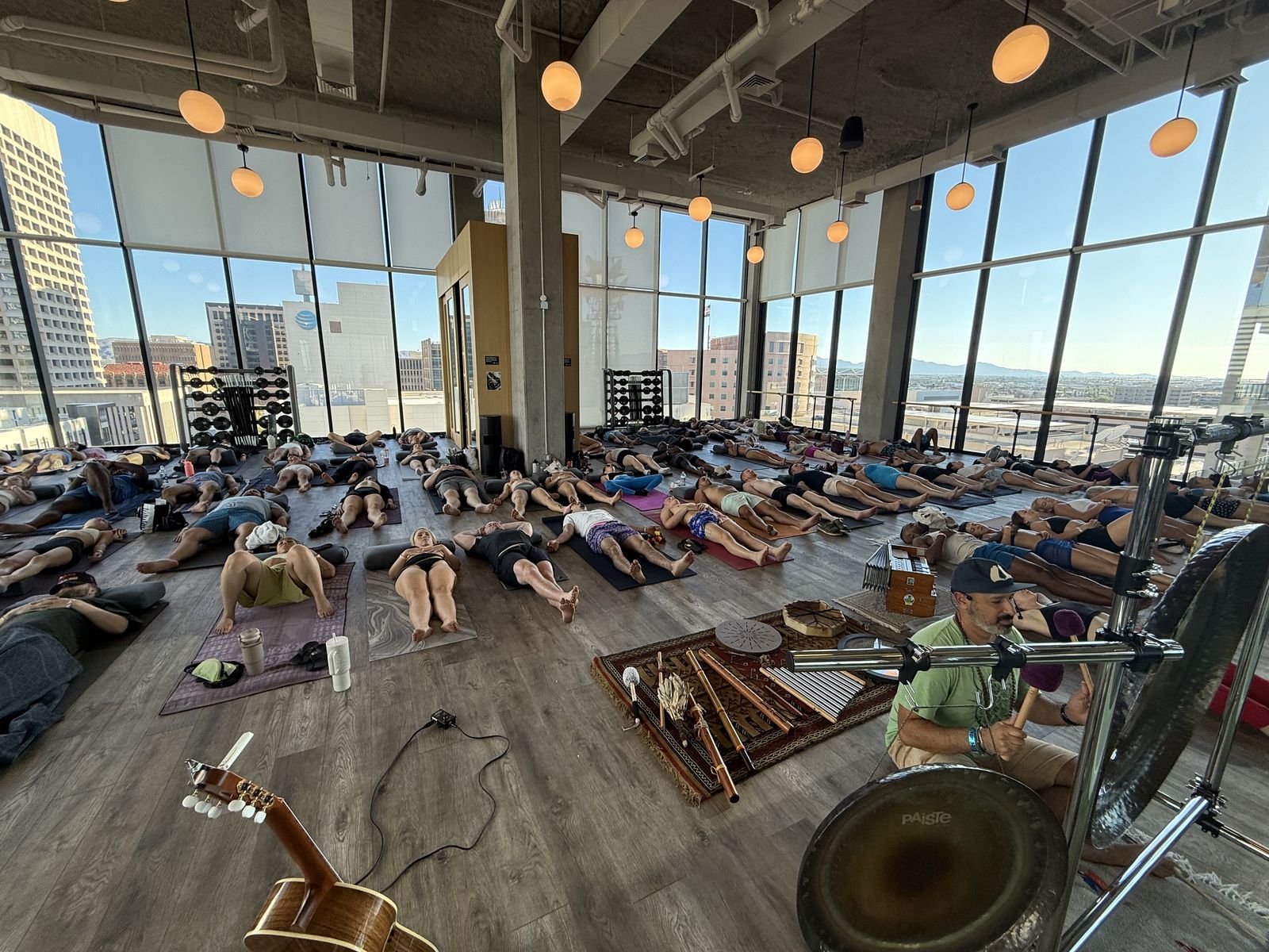 A bright studio with floor to ceiling windows over downtown Phoenix, the floor filled with people lying on mats, Hamid seated at a gong in the foreground