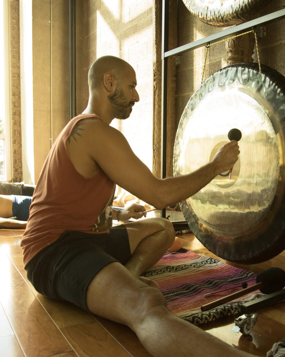 Hamid sitting on a studio floor in front of a large gong, striking it with a soft mallet, with someone lying down behind him in the window light