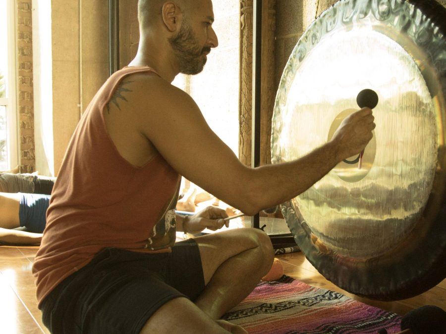 Hamid sitting on a studio floor in front of a large gong, striking it with a soft mallet, with someone lying down behind him in the window light