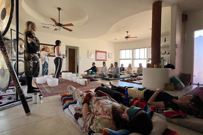 A sound session in progress in a living room at New River, with five people lying down under blankets, a row of students seated behind them and gongs on stands along the wall