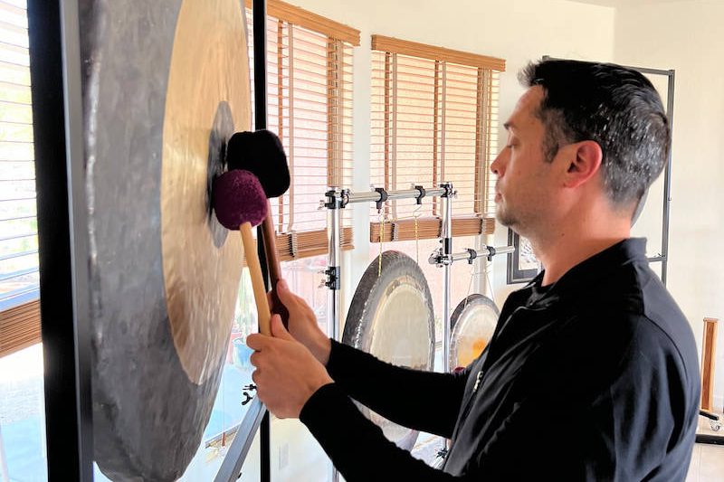 A student at the New River training playing a large hanging gong with a soft mallet, with three more gongs on stands behind