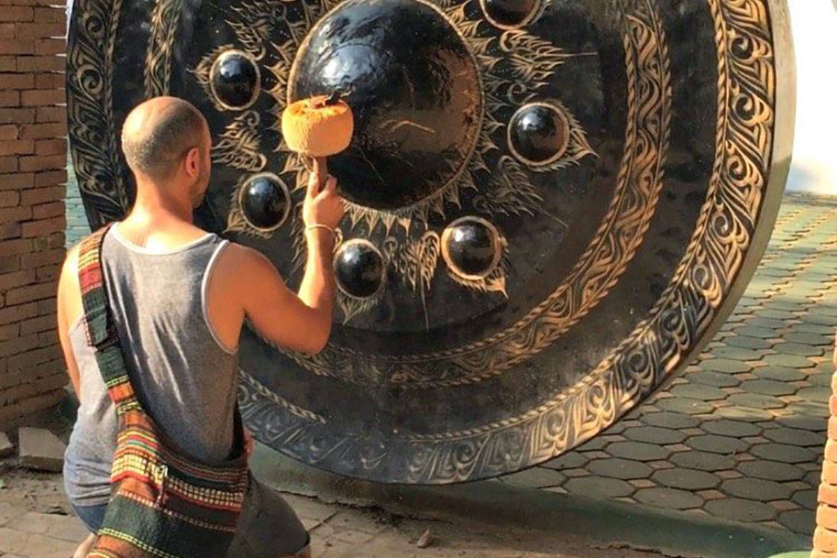 Hamid kneeling to play a large ornate Thai gong hanging outdoors, its face covered in raised bosses and carved detail