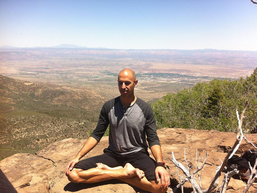 Hamid sitting cross legged on a rock ledge with his eyes closed, a wide Arizona valley opening out below him