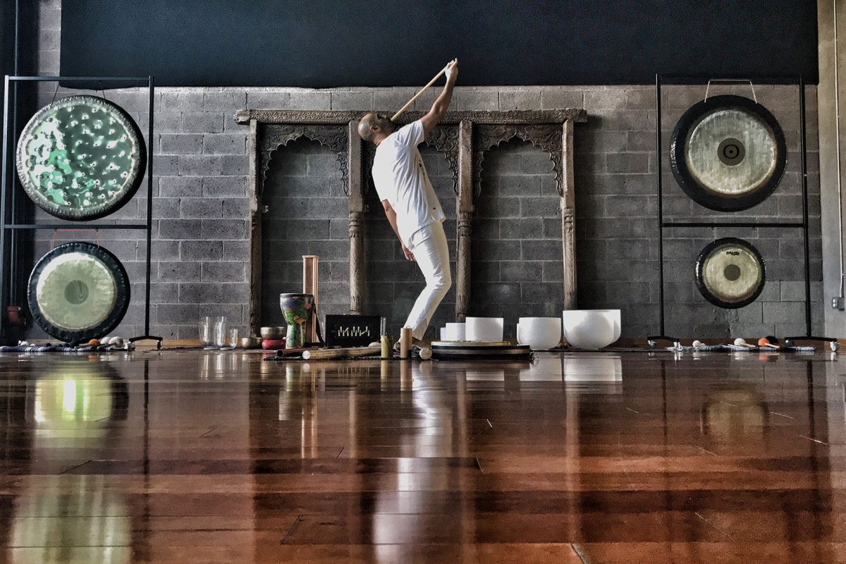 Hamid standing in a studio playing a long overtone flute, with four gongs on stands and a row of crystal bowls along the wall behind him