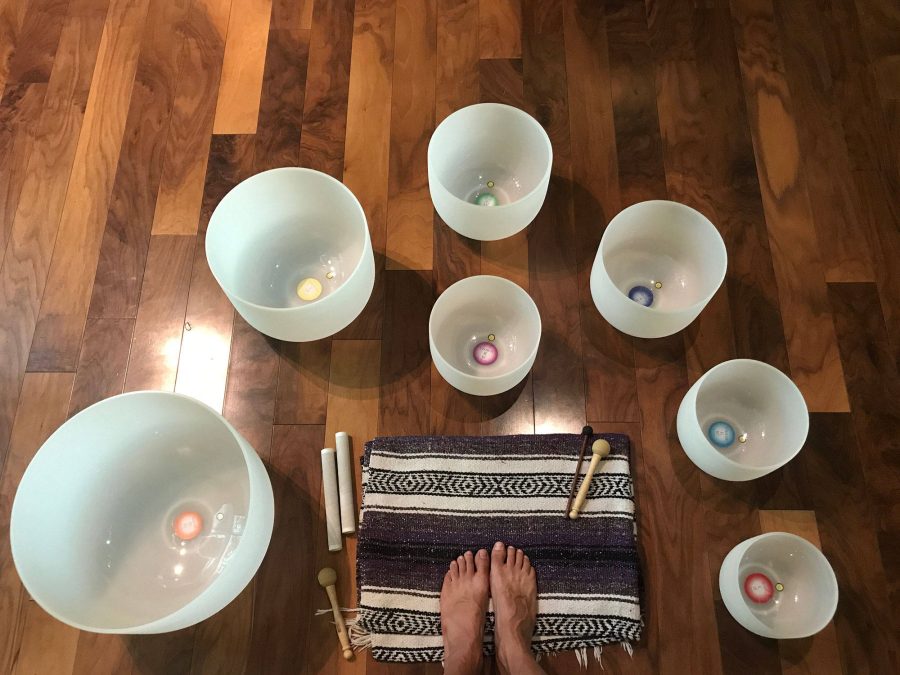 Seven frosted crystal singing bowls arranged in a circle on a wooden floor, each labelled with its note and chakra