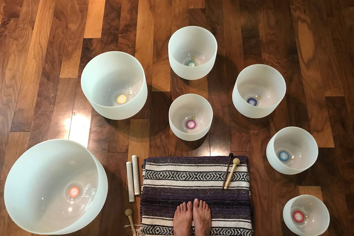 Seven frosted crystal singing bowls arranged in a circle on a wooden floor, each labelled with its note and chakra
