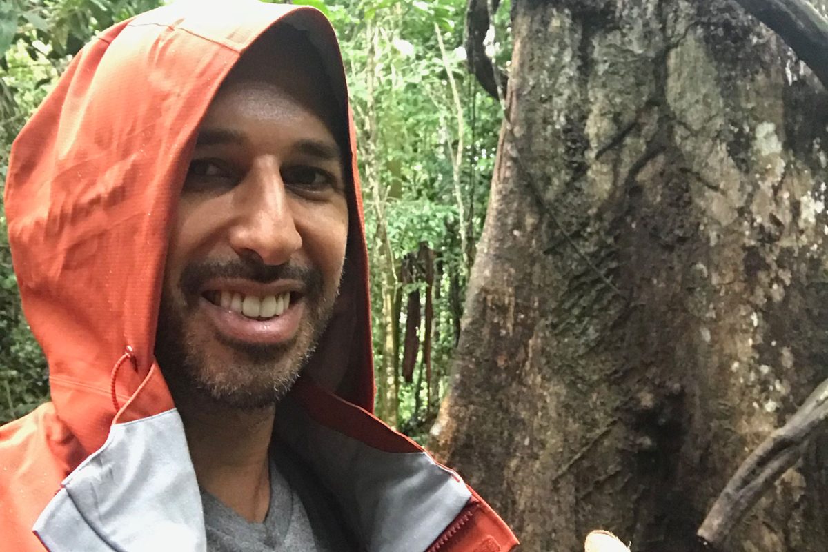 Hamid in a rain jacket beside a large jungle tree, a cut length of ayahuasca vine propped against the trunk
