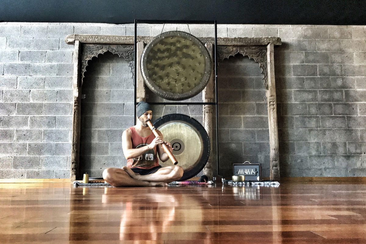 Hamid sitting cross legged on a studio floor playing a flute, two gongs on a carved wooden stand behind him