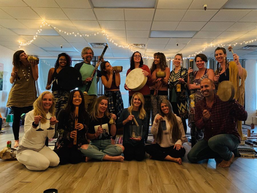 A training group standing and kneeling together in a studio under strings of fairy lights, everybody holding an instrument, singing bowls and flutes and frame drums among them