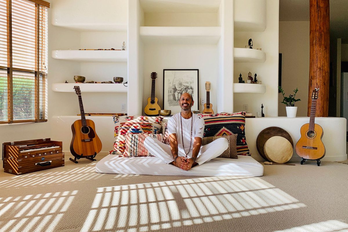 Hamid sitting cross legged in a sunlit room with a harmonium, three guitars, a frame drum and singing bowls around him