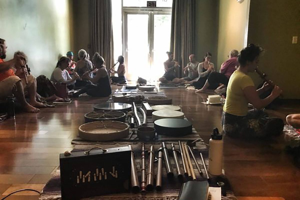 A training group sitting in a circle on a studio floor, each person holding a flute, with a shruti box, frame drums, chimes and mallets laid out on rugs between them