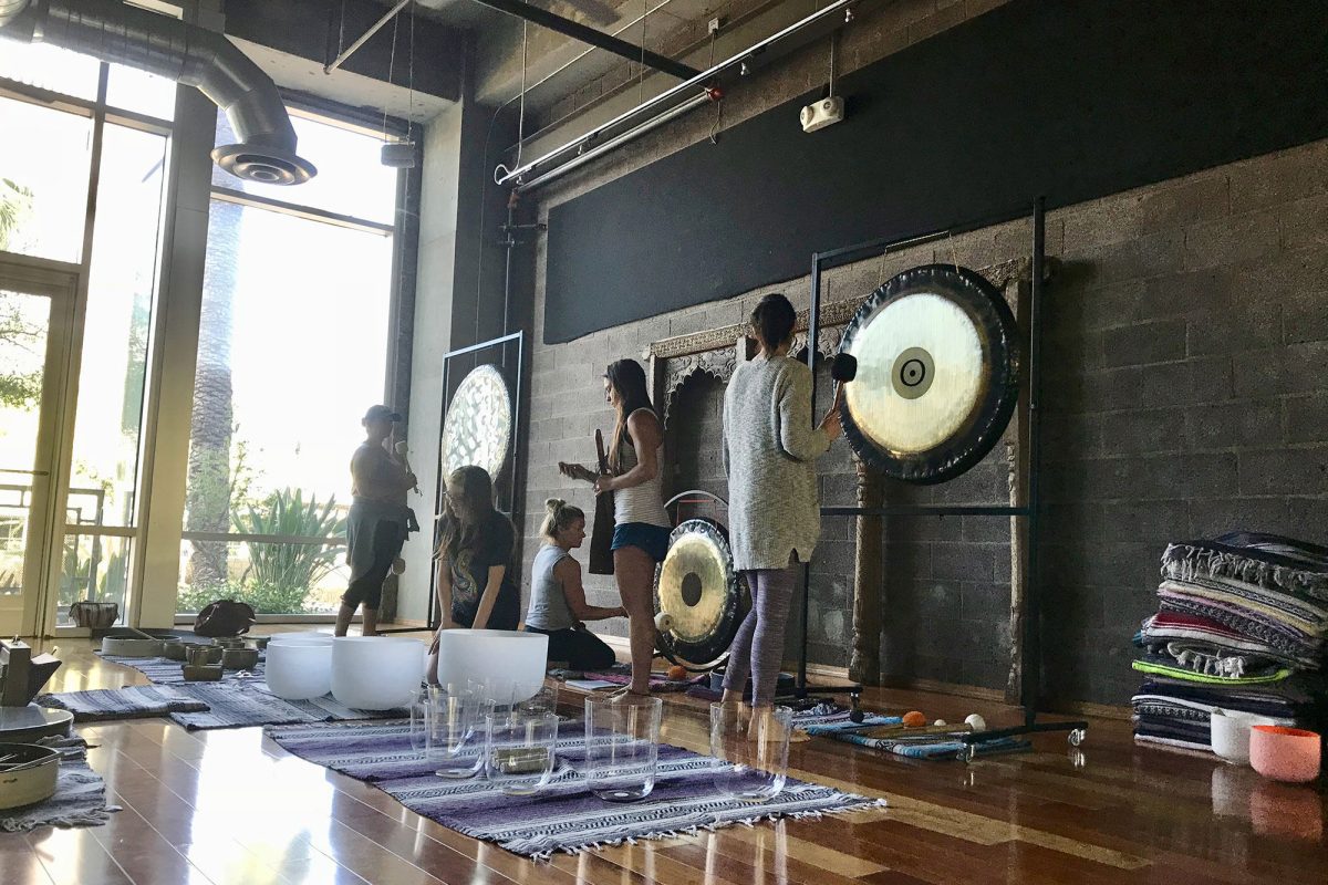 Four students standing at hanging gongs in a studio, playing them with mallets, with crystal bowls and glasses laid out on rugs in front of them