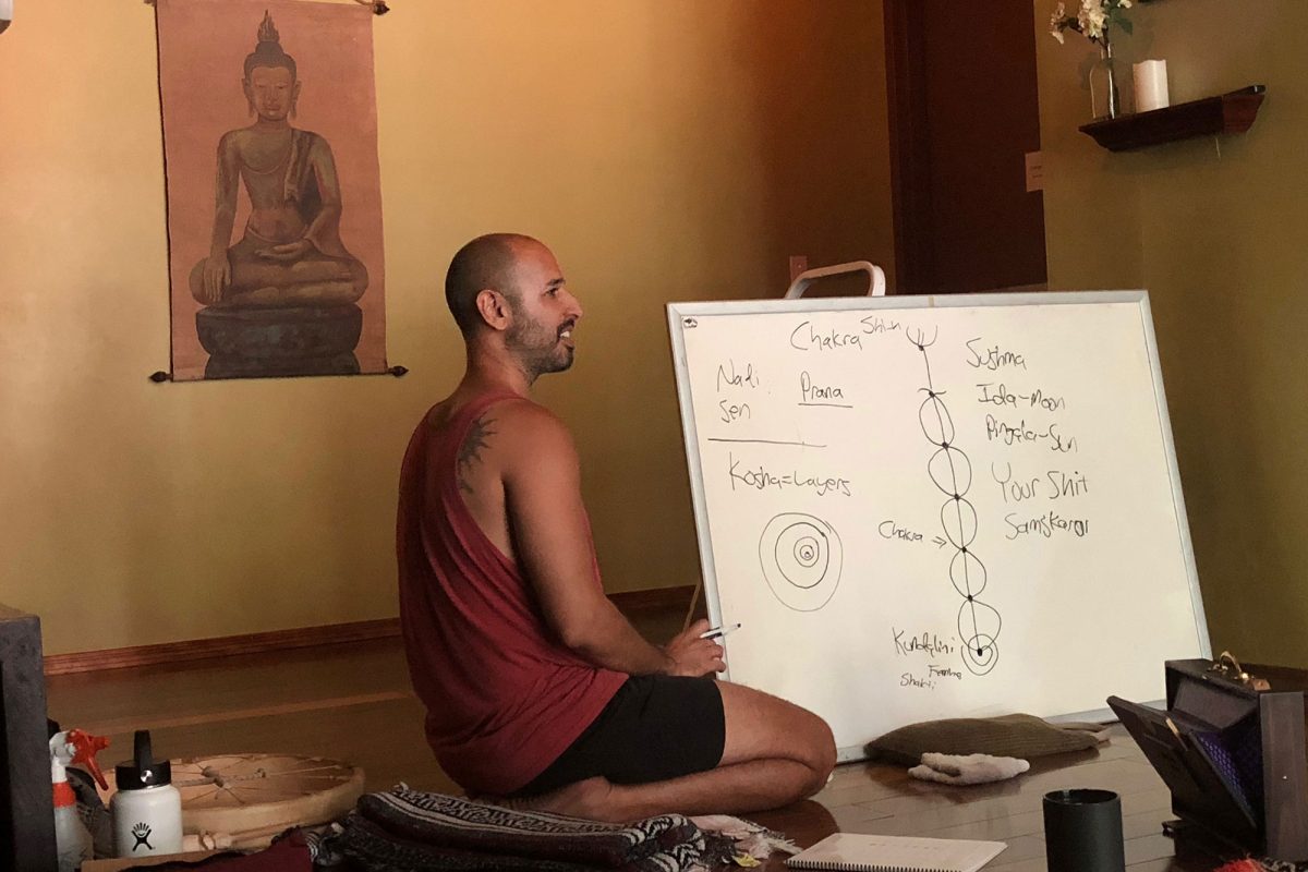 Hamid sitting cross legged on the floor teaching from a handwritten whiteboard, a hanging scroll behind him