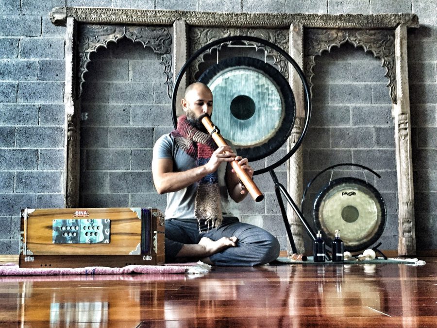 Hamid sitting cross legged on a studio floor playing a long bamboo flute, a hanging gong behind him, a harmonium on the rug beside him and a second gong to his right