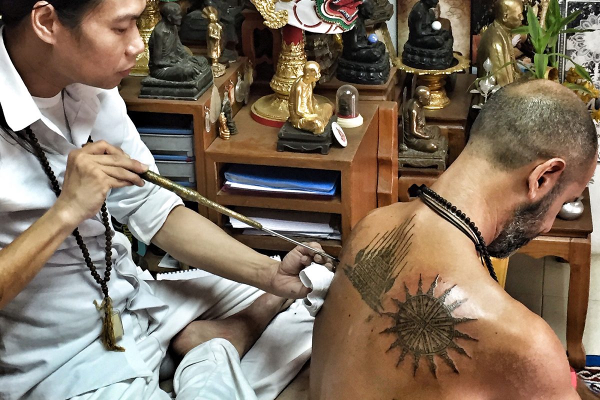 A Thai master tattooing a sak yant on Hamid's back with a traditional rod, in a room full of Buddha figures