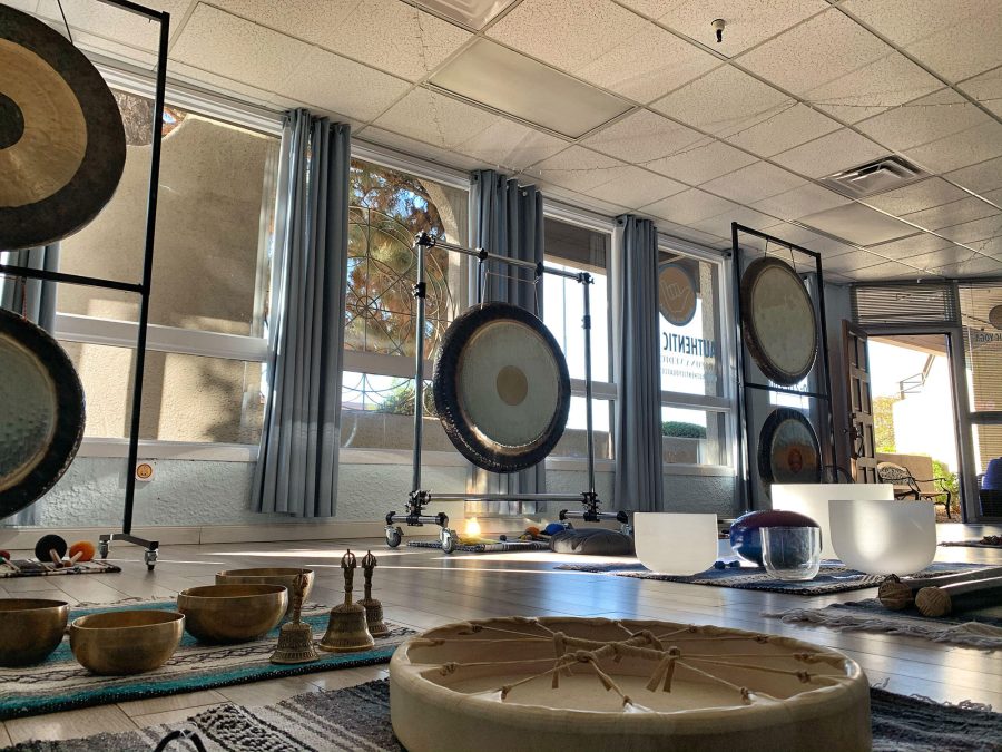 A studio laid out for a training, gongs on stands along a wall of windows, Himalayan bowls, crystal bowls, bells and a frame drum on rugs across the floor