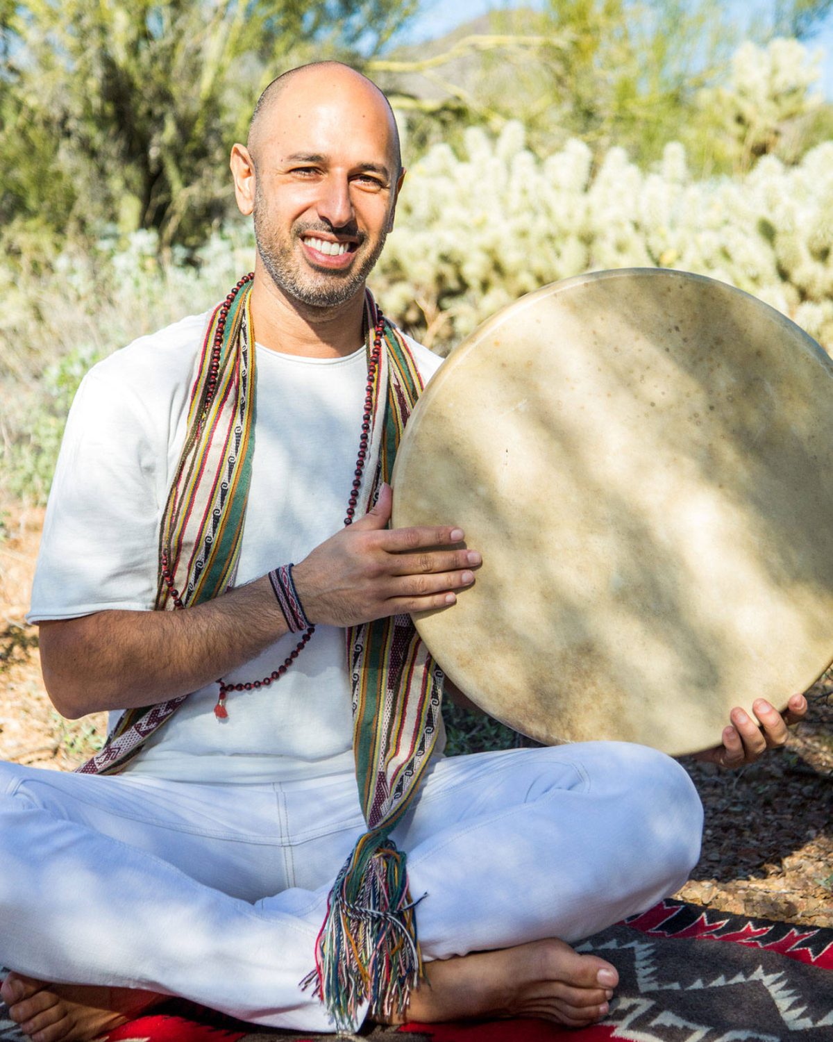 Hamid sitting cross legged outdoors in the desert, smiling, holding a frame drum upright with one hand resting on its skin