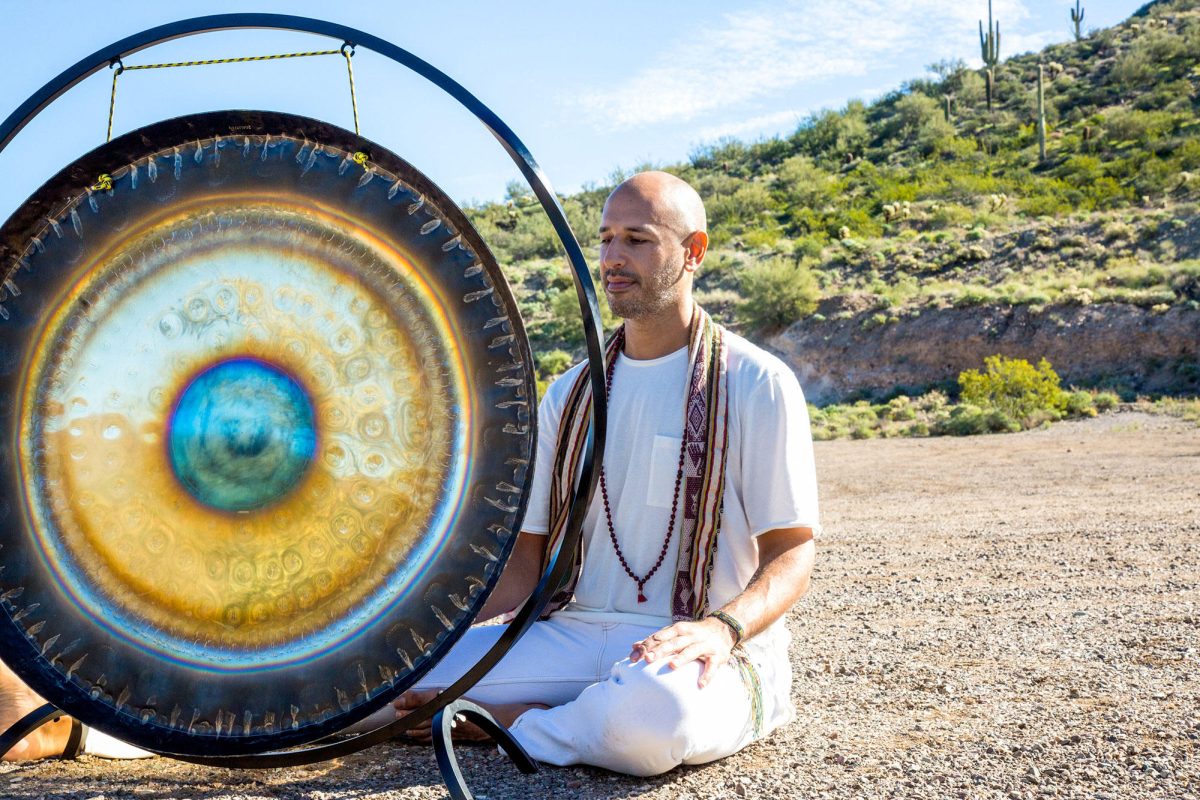 Hamid sitting cross legged on desert ground beside a large gong on a stand, its face burnished gold and blue, saguaro on the hillside behind him