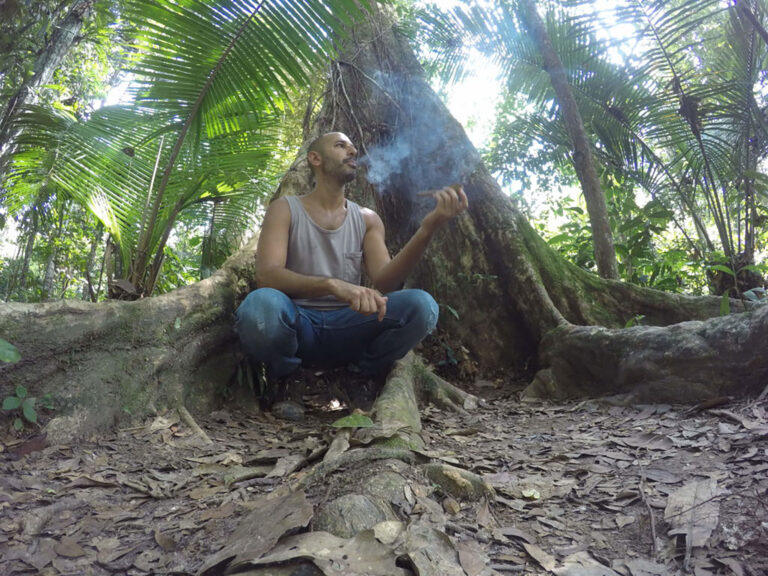 Hamid sitting cross legged in the buttress roots of a large jungle tree in Peru, smoking a mapacho, with smoke rising past his face