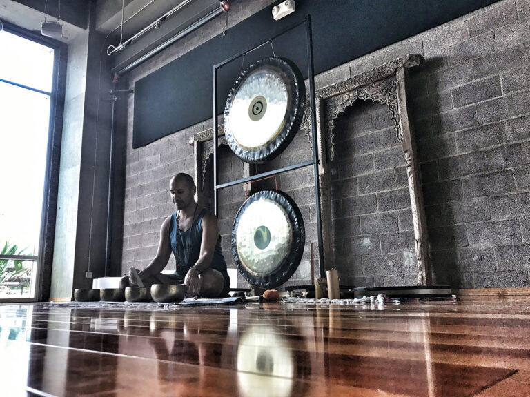 Hamid seated on a studio floor with a row of Himalayan bowls in front of him and two large gongs hanging on a stand beside him
