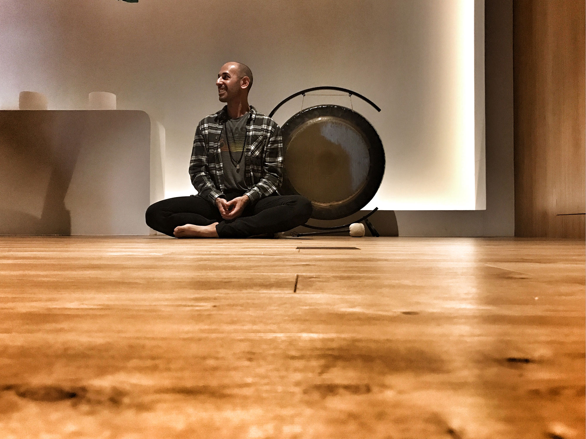 Hamid sitting cross legged on a wooden floor beside a gong on a stand, looking off to one side
