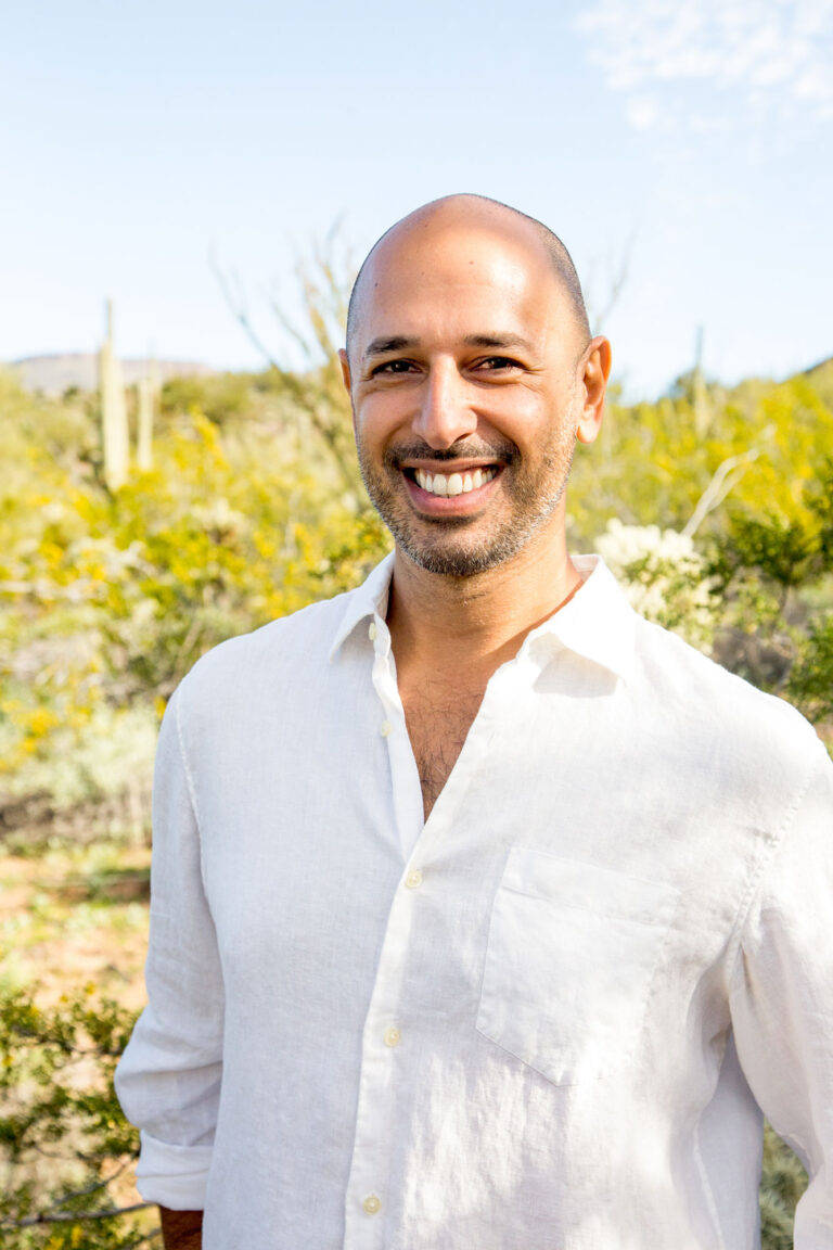 Hamid Jabbar smiling in a white linen shirt, standing in the Sonoran desert with saguaro and blooming palo verde behind him